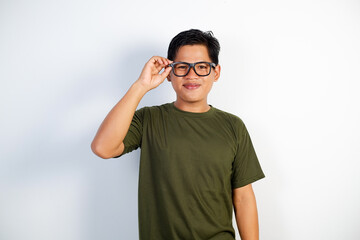 Fototapeta premium A poised young Asian man in a green casual T-shirt adjusts his glasses stands against a white background.