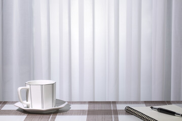 Tea cup, notebook, pen on table, white pleated curtain background. Copy space
