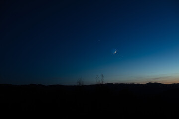 Crescent young Moon with stars, planets and countryside silhouettes.