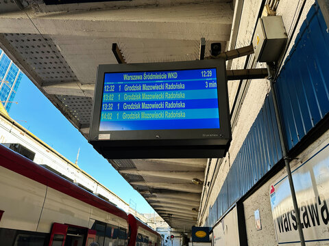 At a Warsaw train station a blue digital departure board displays schedule lines in Poland.
