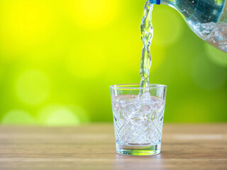 Close-up of clear fresh water being poured into a transparent glass with ice cubes on a wooden table. Natural green bokeh background creates a refreshing, healthy, and clean drinking concept.