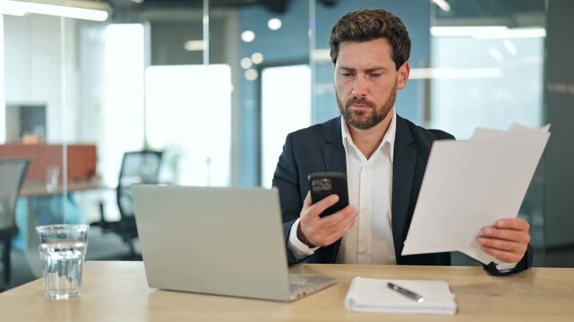 Serious businessman in a dark suit meticulously compares documents with information on a smartphone at a modern office desk, demonstrating focus and eventual satisfaction.