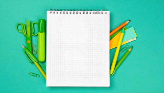 Overhead shot of a spiral notebook on a teal surface with assorted green and orange office and school supplies. White, blank grid-patterned pages