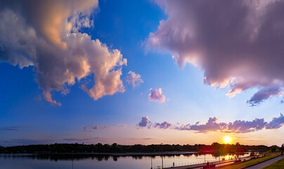 Pink and orange clouds float magically over the river at sunset. Panorama of calm evening fluffy sky near the embankment. Walk before bed, enjoying cloud nature evening. Yellow sun reflection water.