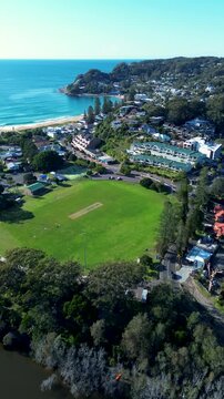 Drone aerial portrait of Avoca Beach local sports fields Heazlett Park with main town shops and cars driving along street with ocean coastline and bushland headland Central Coast tourism Australia