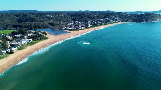 Drone aerial landscape of crystal clear ocean waves breaking on shoreline sandbar at Avoca Beach with river lagoon and residential town housing along bushy coastline headland Central Coast Australia