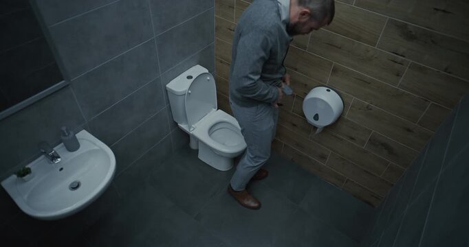 Businessman Taking Off His Pants, Sitting on the Toilet, Suffering from Diarrhea and Indigestion. Man Experiencing Upset Stomach and Digestive Discomfort in Office Restroom During Work. High Angle.
