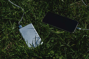 The smartphone charges from an external battery on lush green grass. The elegant design with cables emphasizes the convenience of using outdoor technology, ideal for an eco-friendly theme.
