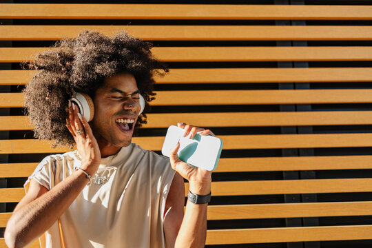 Happy young man enjoying listening to music through headphones and smart phone near striped wall