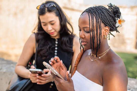 Multiracial women using smartphones outdoors. Two friends standing outdoors checking their phones.