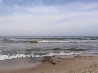 Choppy waves crash onto the sandy shore of the Baltic Sea in Palanga, Lithuania. The sky is partly cloudy, creating a mix of blue and white hues. Dark, wet sand and scattered rocks are visible