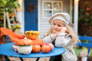 Little smiling girl in beret hat sit at the table in fall cozy garden at home. Child girl relax and dreaming in autumn backyard. Girl play to pumpkins and apples. Thanksgiving Day. Halloween	
