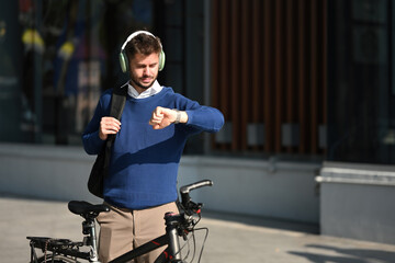 A Caucasian man in the outdoors with a bicycle and a backpack checking the time, Eco-friendly...