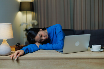 Young Asian woman sleeping at a home office desk with a laptop and coffee cup, experiencing burnout and fatigue from a long night of working late on a project
