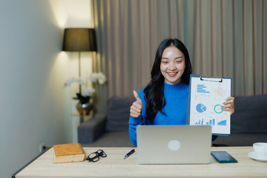 Young Asian woman working from home presents financial charts on a clipboard during a laptop video call, smiling and giving a thumbs up to signal business success and progress