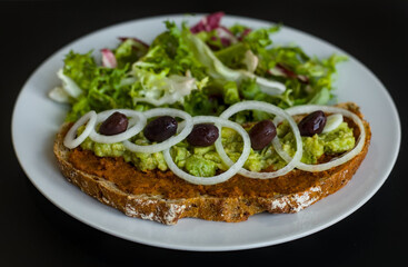 Sourdough Slice of Bread with Pesto, Smashed Avocado, Onions, Black Olives, Mixed Leaf Red Green Salad on a White Plate and Black Background