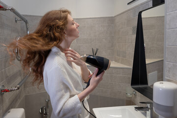 Post-shower self-care: woman drying long red hair with hairdryer near bathroom mirror