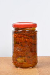 A glass jar filled with preserved sundried tomatoes and olive oil stands on a wooden surface against a white background