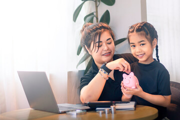 Mother teaches daughter about saving money with a pink piggy bank at home during the day