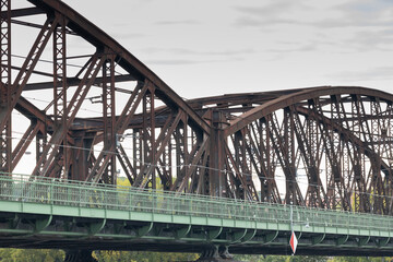 Obraz premium Steel bridge structure with intricate design elements, showcasing rusted metal beams and green railings, spanning over a calm river under a cloudy sky, symbolizing engineering marvel