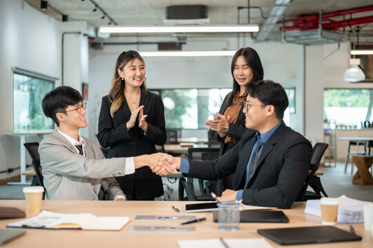 Businesswoman clapping hands while looking at male coworkers shaking hands at wooden table in office - Powered by Adobe