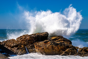 Impressive breaking wave on the granite rocks at Matosinhos beach near Porto in Portugal. Strong surf with spray, splashes, and foam cause constant erosion on the coast on a sunny day with blue skies.