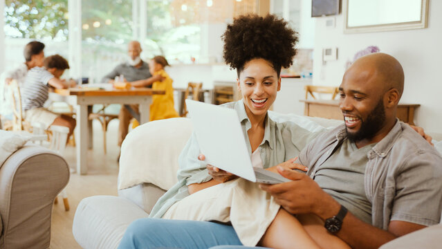 Couple At Home Using Laptop To Shop Or Book Holiday With Multi-Generation Family In Background