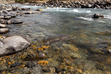 Close up view of clear river with sunlight reflecting on colorful stones and pebbles under the water. The natural ripples and shimmering light patterns create a peaceful and refreshing atmosphere.