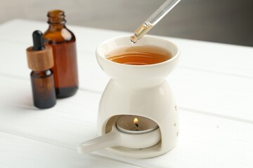 Dripping essential oil from pipette into aroma lamp with burning candle on white wooden table against grey background, closeup