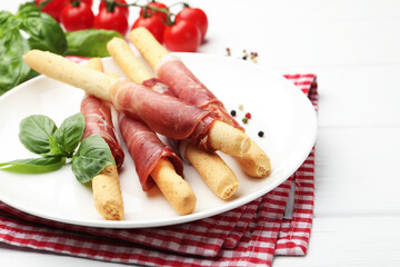 Delicious grissini sticks with prosciutto, peppercorns and basil on white wooden table, closeup