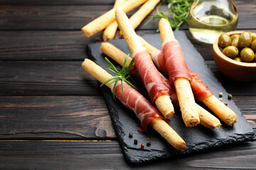 Delicious grissini sticks with prosciutto, peppercorns and rosemary on wooden table, closeup. Space for text