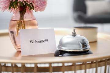 Card with word Welcome, desk bell, candle and vase with flowers on table indoors, closeup
