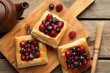 Tasty puff pastries with berries, teapot and brush on wooden table, flat lay
