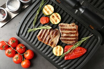 Electric grill with beef steaks, vegetables and rosemary on grey table, flat lay