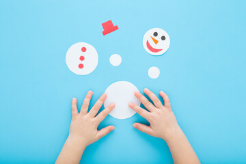 Baby boy hands playing and putting together paper snowman puzzle on blue table background. Pastel color. Flat lay. Homemade jigsaw. Closeup. Point of view shot. Children development. Top down view.