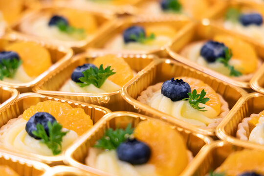 Close-up of assorted mini fruit tarts in golden trays, decorated with blueberries, orange slices. Colorful and elegant dessert presentation, perfect for catering, bakery menus, or food blogs.