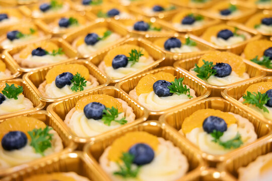 Close-up of assorted mini fruit tarts in golden trays, decorated with blueberries, orange slices. Colorful and elegant dessert presentation, perfect for catering, bakery menus, or food blogs.