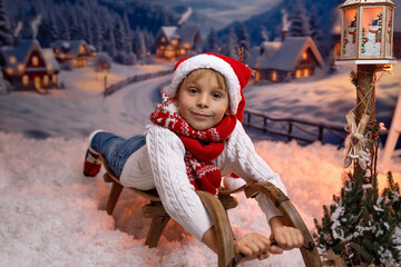 Cute white child, blond boy,, sitting on a sledge in the snow, outdoors, sunset in the mountains