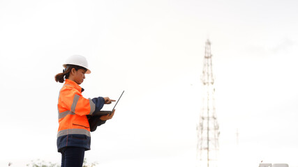 Naklejka premium A woman in bright safety gear stands with a tablet, analyzing information while near a telecommunications tower under a clear sky. She focuses on her task, ensuring safety and efficiency