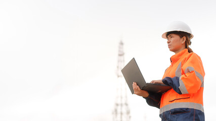 A worker stands outside on a construction site, dressed in bright safety gear and a hard hat, using a laptop to check project details near a tall communication tower