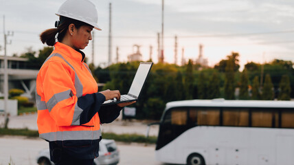 A construction worker dressed in safety gear stands outside at sunset, focused on a laptop. An...