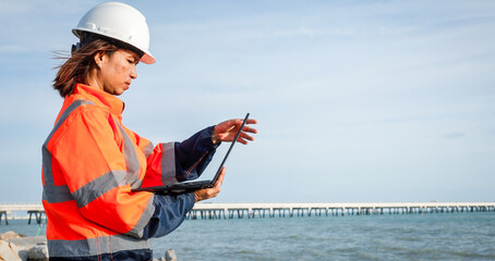A construction worker wearing a hard hat and high-visibility jacket studies plans on a laptop at the water's edge near a dock during a sunny day