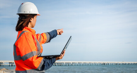A professional woman in safety gear points towards a coastal construction site while using a laptop. The clear sky and water set a calm backdrop for her work