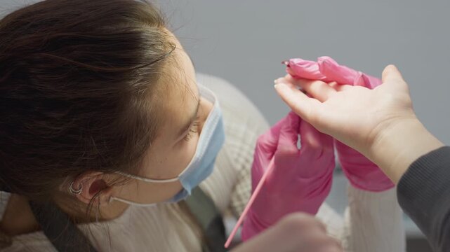 Nail technician wearing face mask and pink gloves bent over while carefully applying hardener to client fingernail using precision brush, visible piercing on ear and focused posture