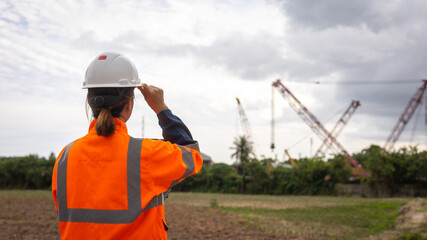 A worker in an orange safety vest and helmet watches cranes at a construction site near a rural area. The sky is cloudy, hinting at possible rain