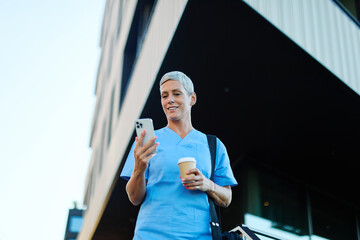 Nurse in scrubs enjoying coffee and using a mobile phone outside a modern medical facility during a...