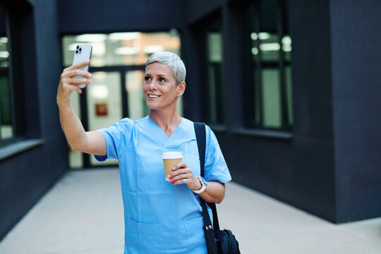 Nurse in scrubs enjoying coffee and using a mobile phone outside a modern medical facility during a sunny day - Powered by Adobe