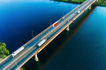 Aerial drone view of bridge asphalt road with cars and cargo trucks driving over blue river or lake water. Logistic and delivery concept.