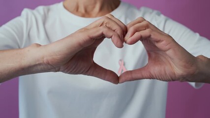 Zoom out shot of anonymous woman making heart shape with hands around breast cancer awareness pink ribbon wearing white t-shirt isolated on pink background