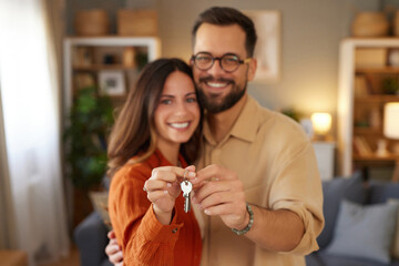 Couple joyfully holds up keys to their new home, smiling and embracing in a warm and inviting living room filled with personal touches and soft lighting.